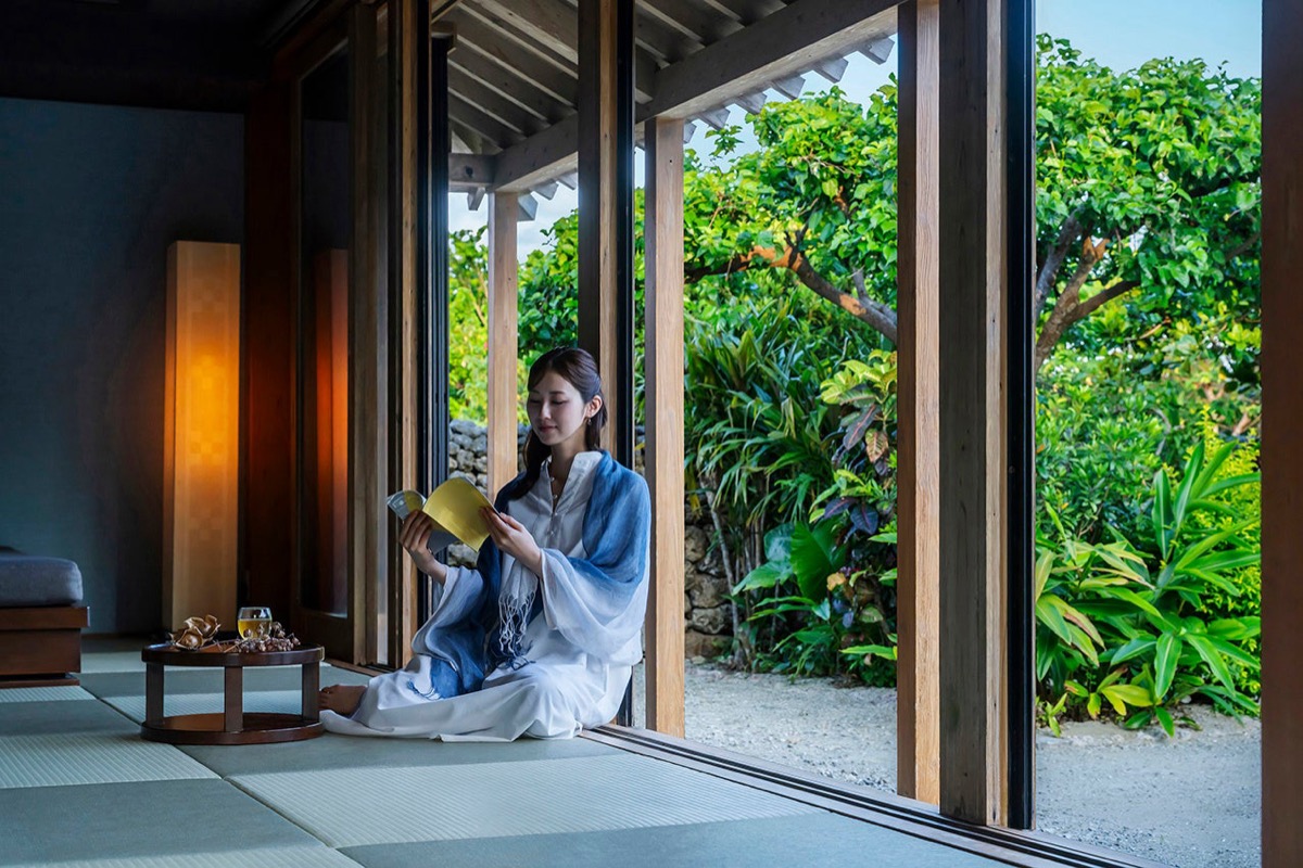 琉球赤瓦の客室の畳の縁側で、白い装いに青いストールを掛けた女性が本を読み、ガラス戸の外に南国の緑の庭が広がる様子