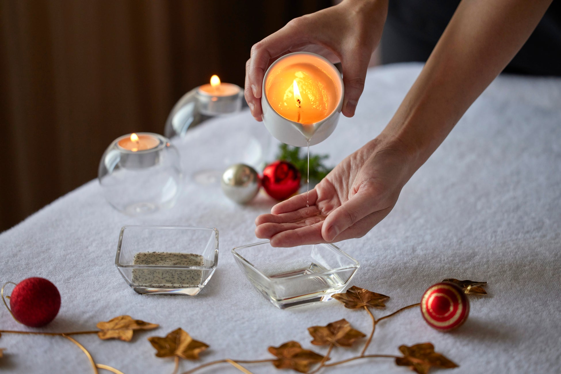 火のともったキャンドルから手のひらにオイルを垂らす様子。台の上にクリスマスオーナメントが並ぶ